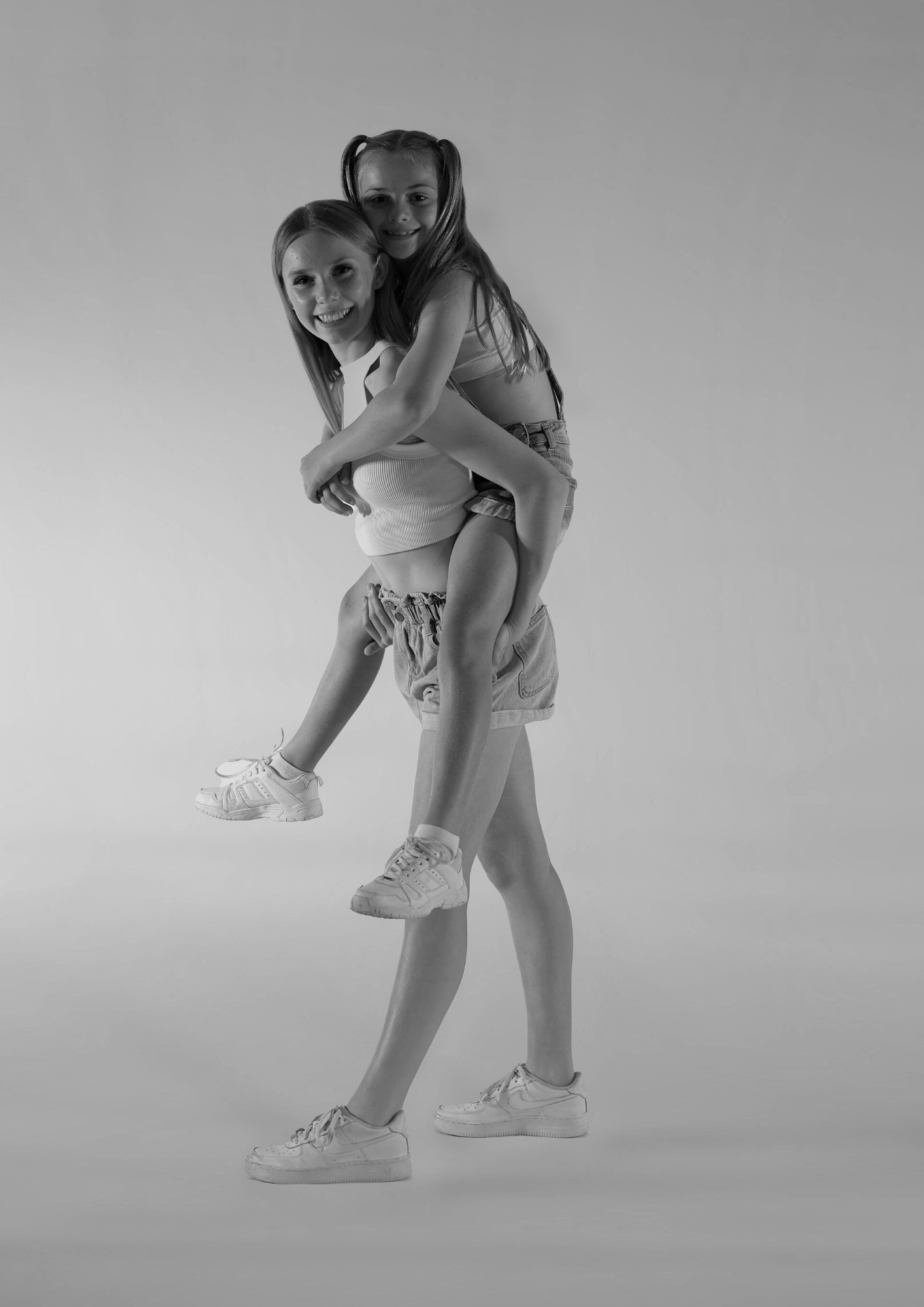 Two young girls smiling at the camera, one giving a piggyback ride at The League Studios in Hervey Bay